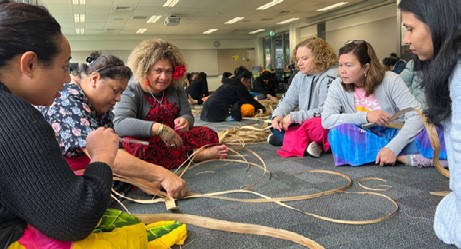 The photo shows six women sitting in a circle on carpet, weaving