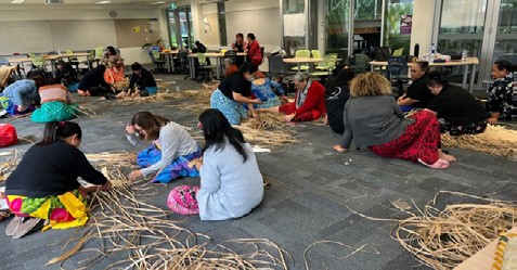 This is a long shot of two dozen people sitting on carpet, weaving