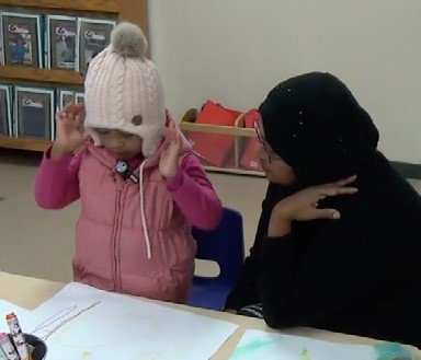 This is a photo of a pre-schooler showing her teacher a drawing and gesturing