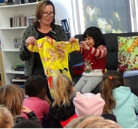 A photo of a teacher holding up a dress in front of the class, while a pre-schooler points out features on the dress