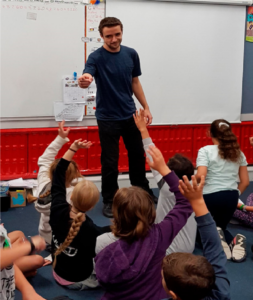 This is a photo of a male teacher with kids on the mat in front of him. Many of the kids have one of their arms raised, as if they want to answer a question the teacher has asked