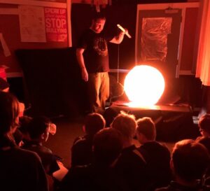 This is a photo of a teacher in front of a class. The room is darkened and there's a bright orb, maybe 40cm wide, in front of the teacher