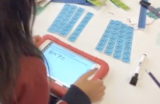 This is a photo of a student sitting at a table holding a tablet. On the table is a pen and some strips of paper