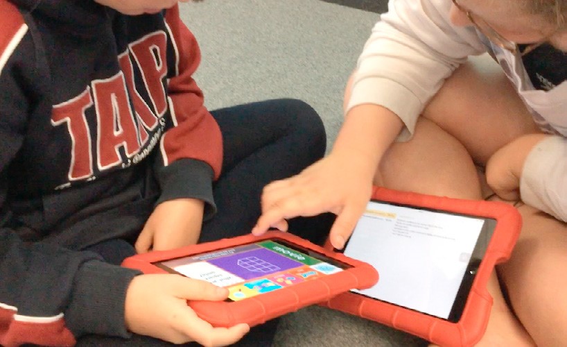 Two students are sitting cross legged on the mat, looking at iPad screens