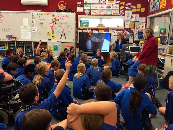 This is a photo of about two dozen students sitting on the floor looking toward the front of the classroom. Two of the children have their arms raised. At the front of the classroom are two teachers, and two students who appear to be showing their presentation