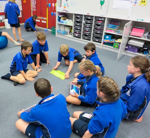 This is a photo of 8 kids sitting on the floor having a discussion. One of them is holding an iPad