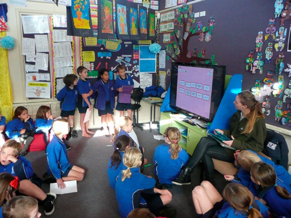 This is a photo of four children standing in front of their class, showing their presentation on a large flat screen TV