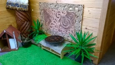 This is a photo of a wide bowl (containing water) sitting on a low platform. There's Māori carvings and plants around the platform