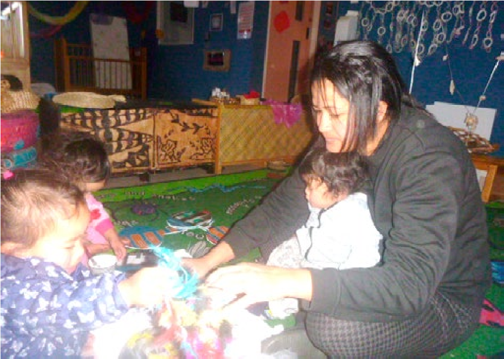 This is a photo of a woman sitting on a mat with a baby in her lap. She is playing with two other children who are sitting close by