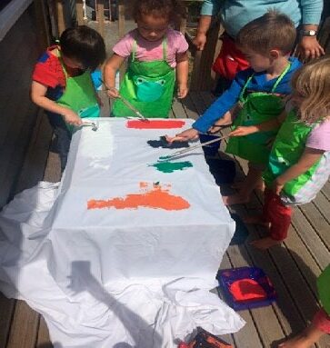 This is a photo of four children around a table, with the partially painted pareu in from of them. The children are holding brushes and are painting