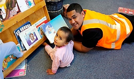 This is a photo of a dad lying face down on the carpet, sharing a book with his child