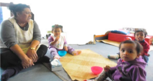 This is a photo of an adult and three children sitting around a woven mat