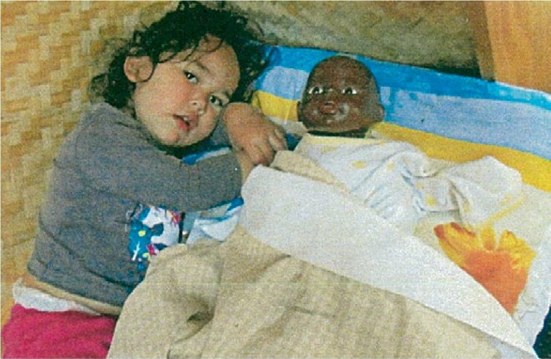 This is a photo of a sleepy looking child lying next to a doll. The doll is lying in a bed on the floor