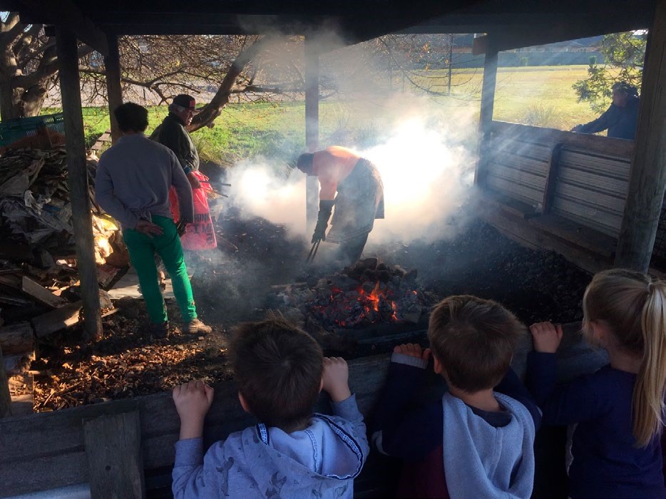 This is a photo of three children watching from behind a fence while 3 people prepare the umu. There's a fire in the middle of the picture, and smoke above the fire