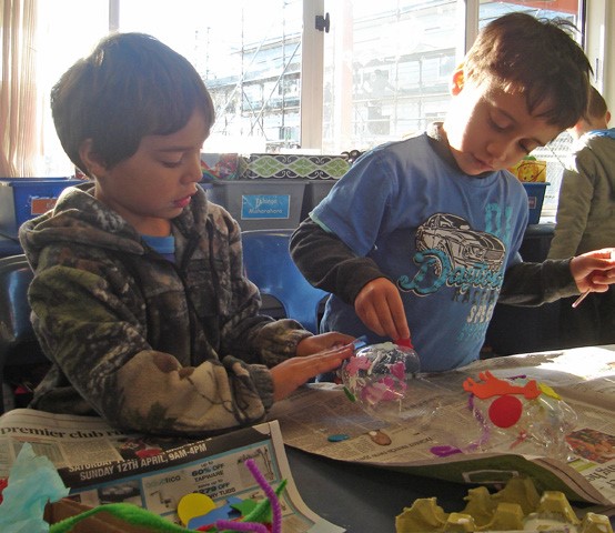 This is a picture of two boys at a school desk. There are recycled materials, such as newspaper and a deconstructed soft-drink bottle, and they appear to be crafting something