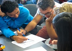 This is a photo of a lecturer and some students at a desk looking down at a piece of paper. They may be trying to work out a solution to a question