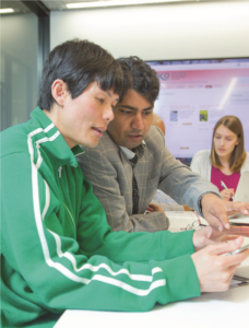 A photo of a man sitting at a table, in discussion with another man standing beside him. They may both be looking at a calculator