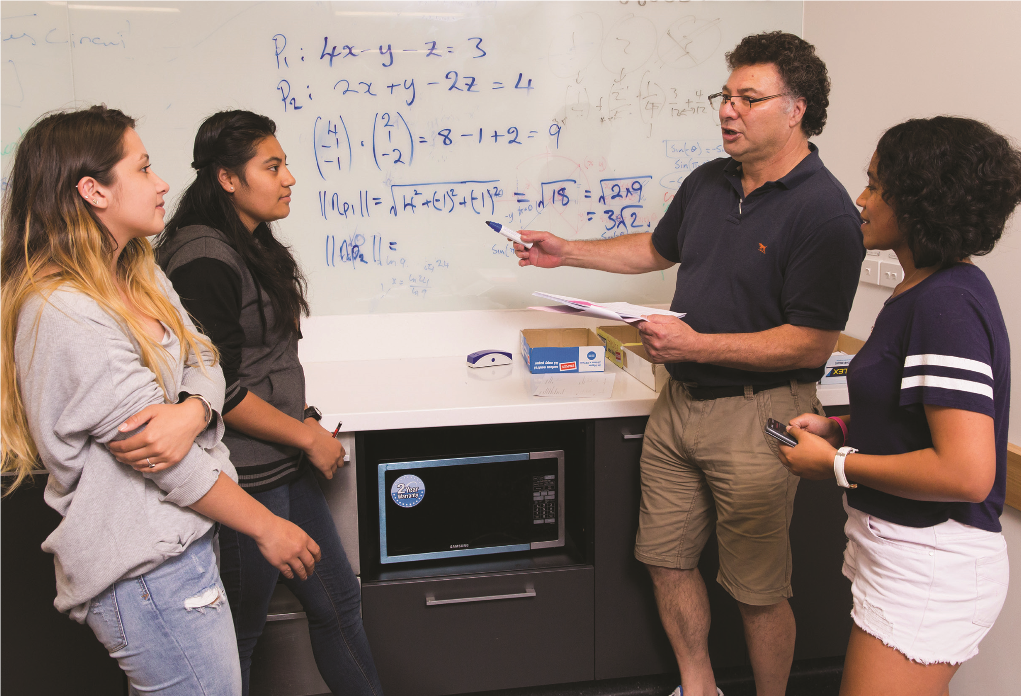 A photo of a lecturer pointing to a whiteboard, discussion an equation with three nearby students