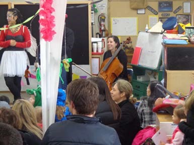 An adult is sitting in front of a cello, while others are gathered around