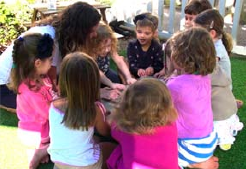 Eight children and teacher sitting in a circle outside, focusing on what could be a musical instrument in the middle of the circle