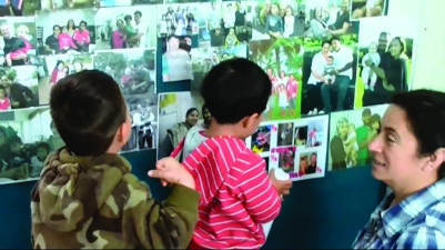 This is a picture of a teacher discussing photos with two children. The pictures are pinned to a board on the wall