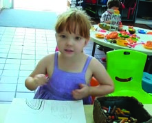 This is a picture of a girl looking at the camera, while her drawing sits on the table in front of her