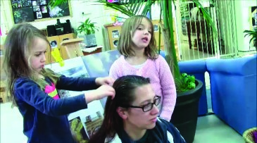 A photo of a teacher sitting, with two girls standing. One of the girls is touching the teacher's hair