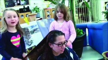 Another photo of a teacher sitting, with two girls standing. One of the girls is touching the teacher's hair