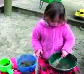 A photo of a child playing with bowls and a spoon in the sandpit