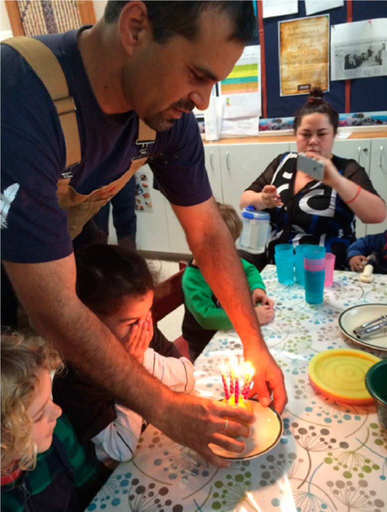 A father is placing a birthday cake on a table in front of some pre-schoolers