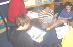 People reading books. The teacher is sitting on a chair, and the students are on the mat