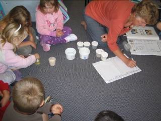 A teacher and half a dozen kids are sitting or crouching on the floor. The kids have containers in front of them. The teacher is writing in an exercise book