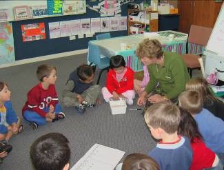 The teacher and children are sitting in a circle. They're concentrating on a container in front of one of the children