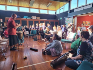 This is a photo of a teacher leading a drum lesson in from of 20 people. He is standing. Many others are sitting on the floor with their drums